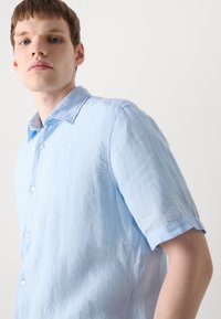Light blue linen short-sleeve shirt with a button-down collar, textured fabric, and visible stitching details on cuffs and placket.