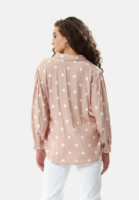 Pink blouse with white polka dots, long sleeves, buttoned cuffs, and a relaxed fit. Soft fabric with slight sheen and subtle pleats at shoulders.