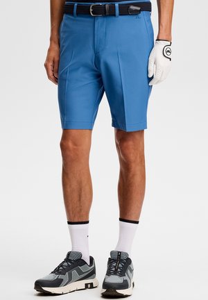 Mann trägt blaue Golfsport-Shorts, schwarzen Gürtel, weißen Golfhandschuh, weiße Socken und schwarz-graue Sportschuhe, steht vor einem einfarbigen Hintergrund.
