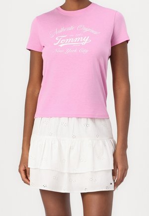 Kvinde iført en lyserød T-shirt med korte ærmer med teksten "Tommy" og en hvid lagdelt nederdel med hulmønster, stående mod en ensfarvet baggrund.