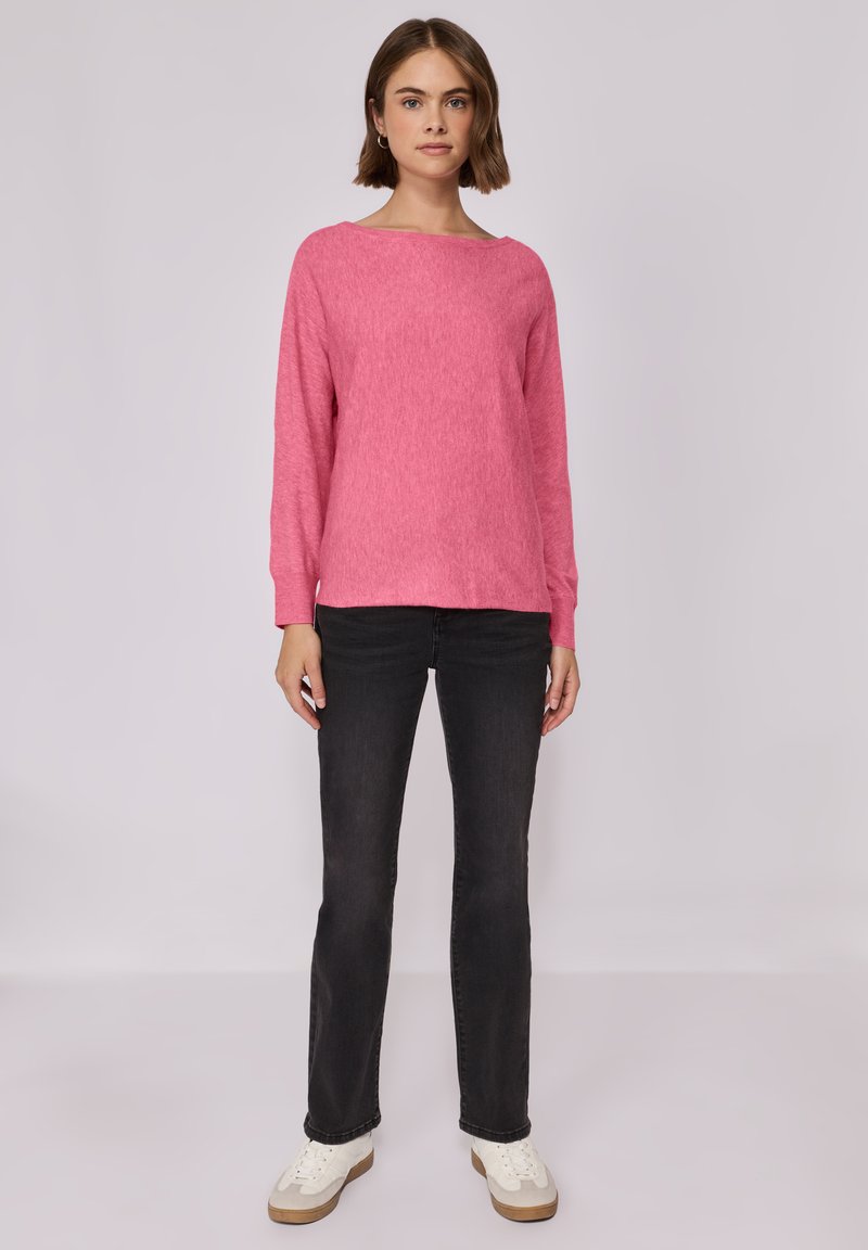 Maglione rosa a maniche lunghe con scollo a barca, abbinato a jeans neri a gamba dritta e sneakers bianche, in piedi su uno sfondo semplice.