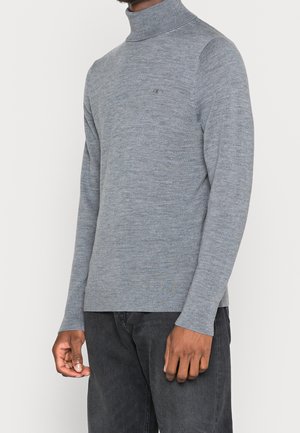 Homme portant un pull à col roulé gris ajusté à manches longues et un pantalon gris foncé, debout devant un fond uni.