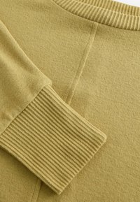Sweat-shirt en vert olive avec un col et des poignets côtelés. Tissu doux et texturé avec des détails de couture subtils au dos.