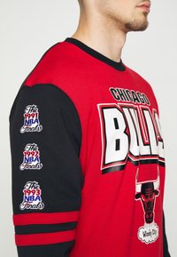 Chemise à manches longues rouge et noire avec le logo des "Chicago Bulls", un graphique de taureau et des années de finales brodées sur les manches. Accents rayés.