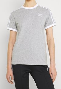 Šedé bavlněné tričko s bílým žebrovaným límcem a manžetami, se třemi bílými pruhy na ramenou a malým logem na hrudi.