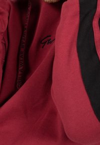 Bordowy sweter z czarnymi akcentami i teksturowanym materiałem. Posiada haftowane logo i dekolt ze sznurkiem. Zbliżenie na materiały.