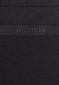 Czarna dzianina o gładkiej fakturze. Posiada czarny pasek z wytłoczonym napisem "HILFIGER" na środku. Prosty wzór, bez dodatkowych motywów.