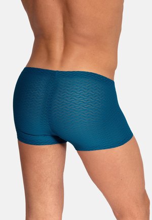 Mężczyzna noszący dopasowane niebieskie bokserki o fakturze, widoczny od tyłu od talii do połowy uda, na jednolitym jasnym tle.