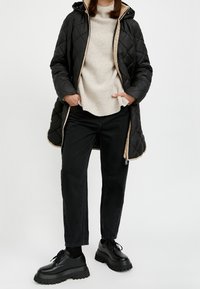 Sort quiltet jakke med beige detaljer, lange ærmer og hætte; kombineret med en beige ribstrikket sweater, sorte jeans og kraftige sorte sko.