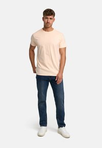 Jasnopomarańczowa bawełniana koszulka z okrągłym dekoltem i krótkimi rękawami, zestawiona z niebieskimi dżinsami i białymi trampkami. Prosty design, bez widocznych wzorów.