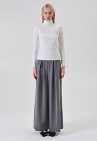 Donna che indossa un maglione a collo alto bianco testurizzato, pantaloni grigi a gamba larga e scarpe nere con punta e fiocchi, in piedi contro uno sfondo neutro.