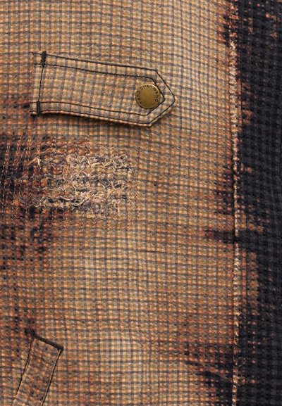 Κοντινή λήψη καφέ καρό υφάσματος με μια ραμμένη επιδιόρθωση και ένα μικρό σκεπαστικό που στερεώνεται με ένα ορειχάλκινο κουμπί με την επιγραφή «WANG».