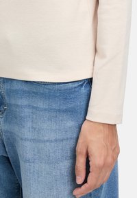 Helles beiges Langarmhemd mit weicher Textur und geradem Saum, kombiniert mit hellblauen Jeans; die Hand ruht auf dem Stoff.