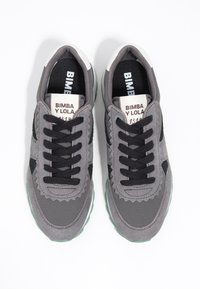 Graue Sneakers mit schwarzen Akzenten, Obermaterial aus Wildleder und Textil, Schnürdesign, gewellte Details und eine hellgrüne Gummisohle.