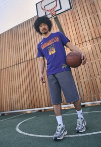 Violets basketbola krekls ar "NUGGETS 15" zelta krāsā, džinsus šorti un pelēki sporta apavi. Ir redzama koka fona un basketbola grozs.