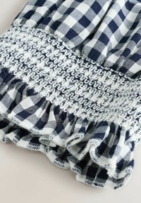 Tissu en vichy bleu marine et blanc avec smocks froncés et détail de bordure volantée sur fond clair.