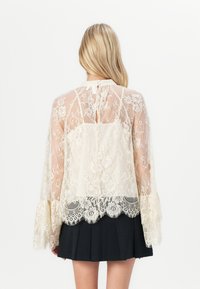 Blusa in pizzo con un design trasparente caratterizzato da motivi floreali e maniche a campana, rifinita con un orlo in pizzo scallop. Colore crema.