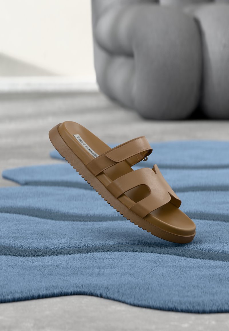 Sandales tongs marron avec une tige synthétique lisse, deux larges brides, une sangle entre les orteils et une semelle en caoutchouc texturée, posées sur un tapis bleu.