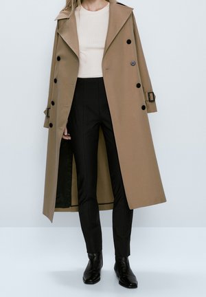 Frau trägt einen beige doppelreihigen Trenchcoat über einem weißen gerippten Top, schwarze maßgeschneiderte Hosen und schwarze knöchelhohe Stiefel vor einem einfarbigen Hintergrund.