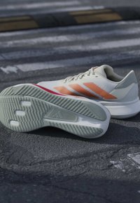 Paire de chaussures de course blanches avec des rayures orange, dont une montre une semelle texturée, posée sur une surface asphaltée près d’un passage piétons.