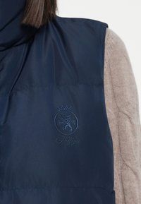 Mornarska puffer prsluk s blagim sjajem, s utisnutim logotipom lava i krune. Nosite ga preko svijetlosmeđeg teksturiranog pulovera.