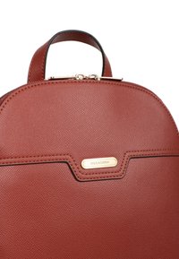 Brązowy plecak ze skóry o teksturowanej powierzchni z zaokrąglonym dnem, z akcentami w złotym kolorze i czarnymi detalami na brzegach. Zamek błyskawiczny na górze.