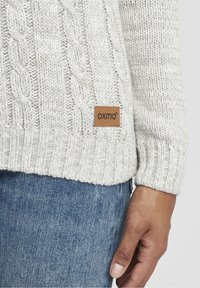 Pull en maille torsadée en mélange de laine gris clair avec ourlet côtelé. Comprend une étiquette de marque en cuir camel. Associé à un jean en denim bleu.