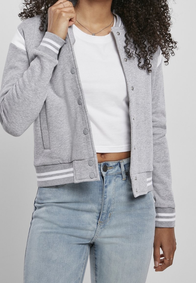 Femme portant une veste varsity gris clair sur un crop top blanc et un jean bleu clair, tenant le col de la veste avec cheveux bouclés visibles.