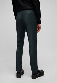 Maßgeschneiderte grüne Hosen mit glatter Textur, zwei Gesäßtaschen und schwarzen Knopfdetails. Getragen mit schwarzen Schuhen, von hinten betrachtet.