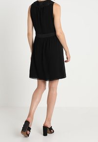 Černé bezrukávové šaty s krajkovým výstřihem, přiléhavým pasem a splývavou sukní. Nosené s černými sandály na blokovém podpatku.