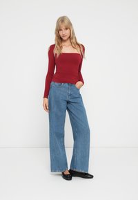 Giovane donna in piedi con una mano in tasca, indossa un fitted top rosso a maniche lunghe, jeans blu a gamba larga e ballerine nere.