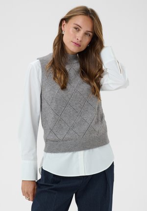 Femme portant un pull sans manches gris en maille sur une chemise blanche à manches longues, avec un pantalon foncé à fines rayures, posant devant un fond neutre.