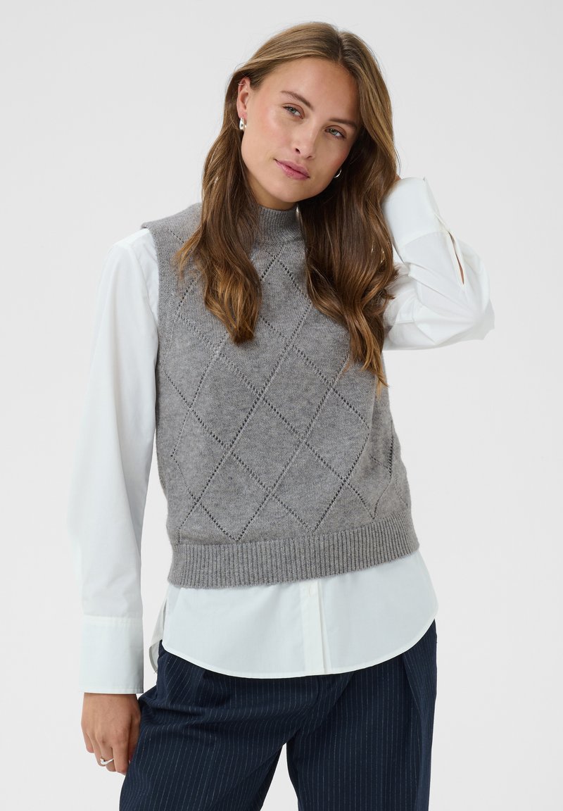 Femme portant un pull sans manches gris en maille sur une chemise blanche à manches longues, avec un pantalon foncé à fines rayures, posant devant un fond neutre.