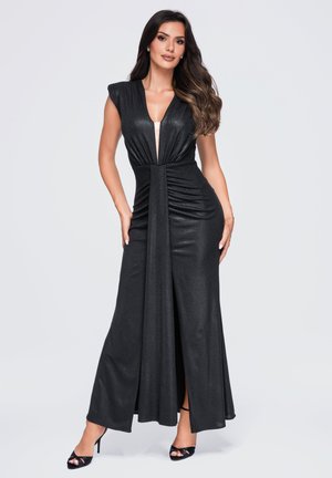 Femme portant une robe de soirée noire sans manches avec un décolleté en V profond et une fente à l'avant, posant sur un fond clair uni.