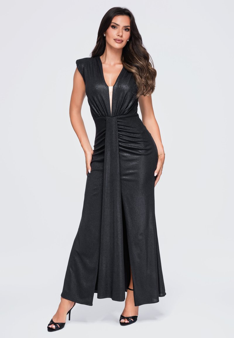 Femme portant une robe de soirée noire sans manches avec un décolleté en V profond et une fente à l'avant, posant sur un fond clair uni.