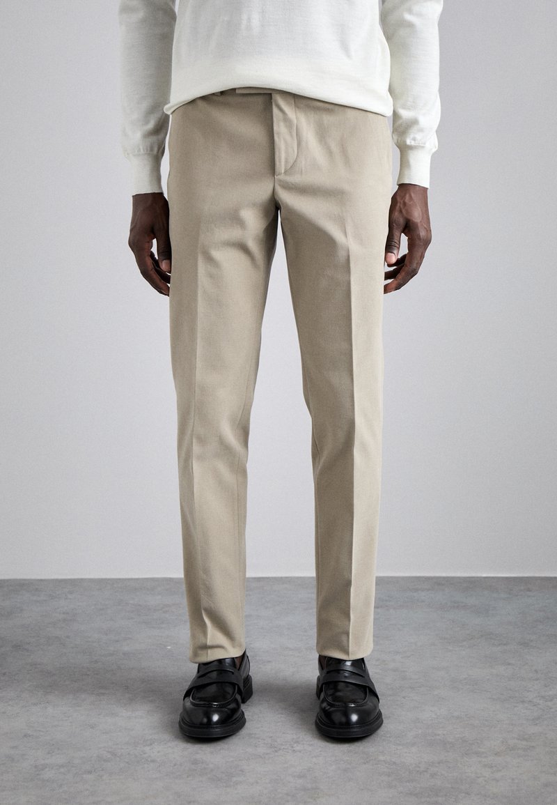Beige maßgeschneiderte Hose mit einer glatten Textur, schlanker Passform und einem Bund, kombiniert mit schwarzen eleganten Schuhen. Der Hintergrund ist neutral.