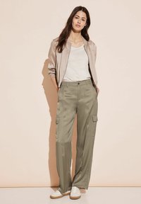 Ljusbeige bomberjacka över en vit topp, tillsammans med olivgröna vida cargobyxor med sidofickor och en slät textur. Vita sneakers.