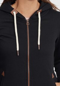 Černá mikina s kapucí na plný zip, krémová šňůrka, bronzové kovové prvky a hnědé detaily na kapsách, s hladkým povrchem.