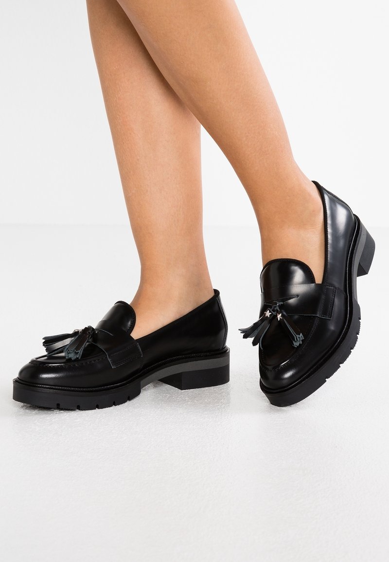 Mocassins en cuir noir avec bout rond, accents à franges et une semelle épaisse et texturée. Fini lisse avec des détails en métal contrastants.