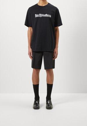 Homme portant un t-shirt noir oversize avec un texte graphique blanc, un short noir longueur genou, des chaussettes noires et des mocassins en cuir noirs.