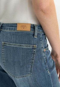 Jean en denim bleu moyen avec une patch en cuir au dos portant l'inscription « Le temps des cerises ». Présente un design distinctif de poche arrière.