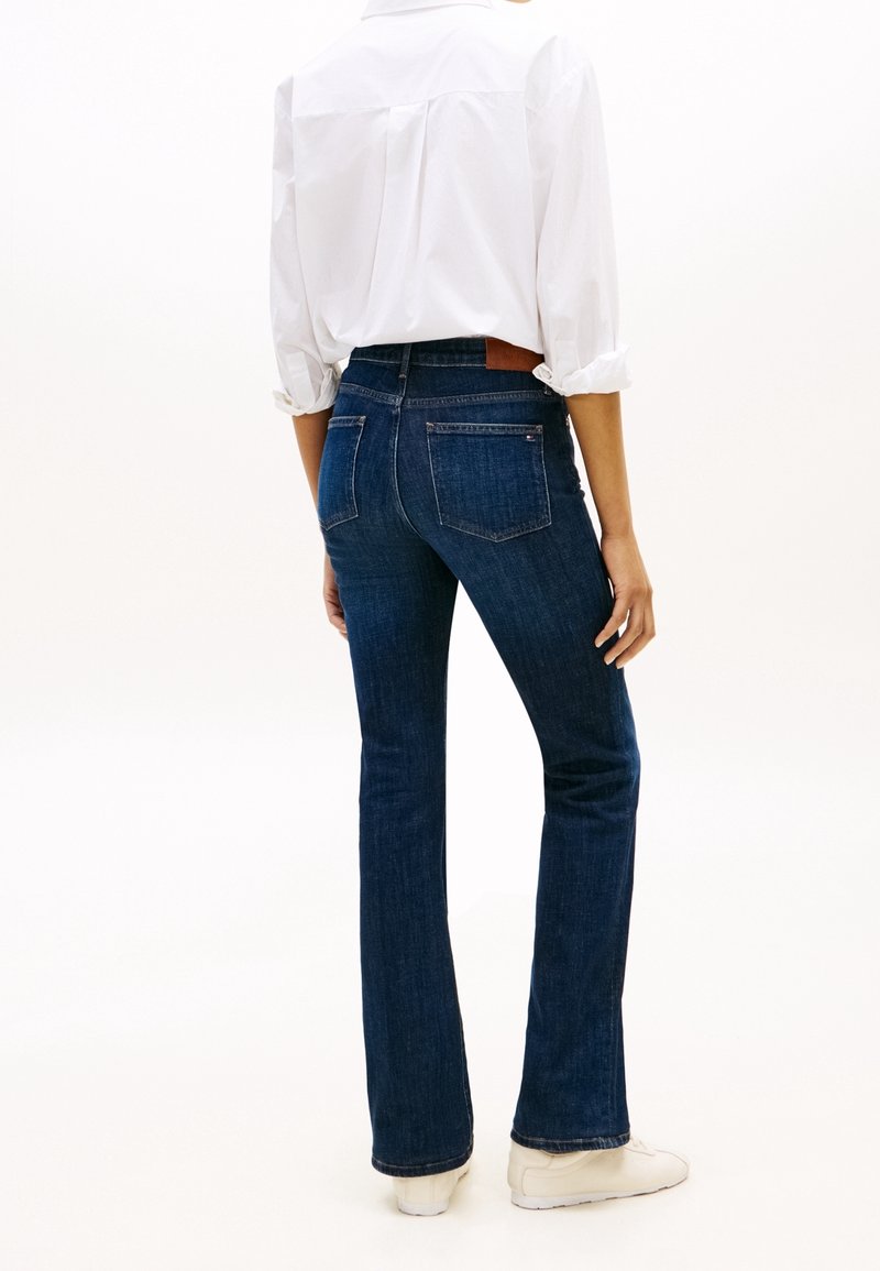 Personne portant un jean bootcut bleu foncé, une chemise blanche à manches longues avec les manches retroussées, et des baskets blanches, vue de dos.