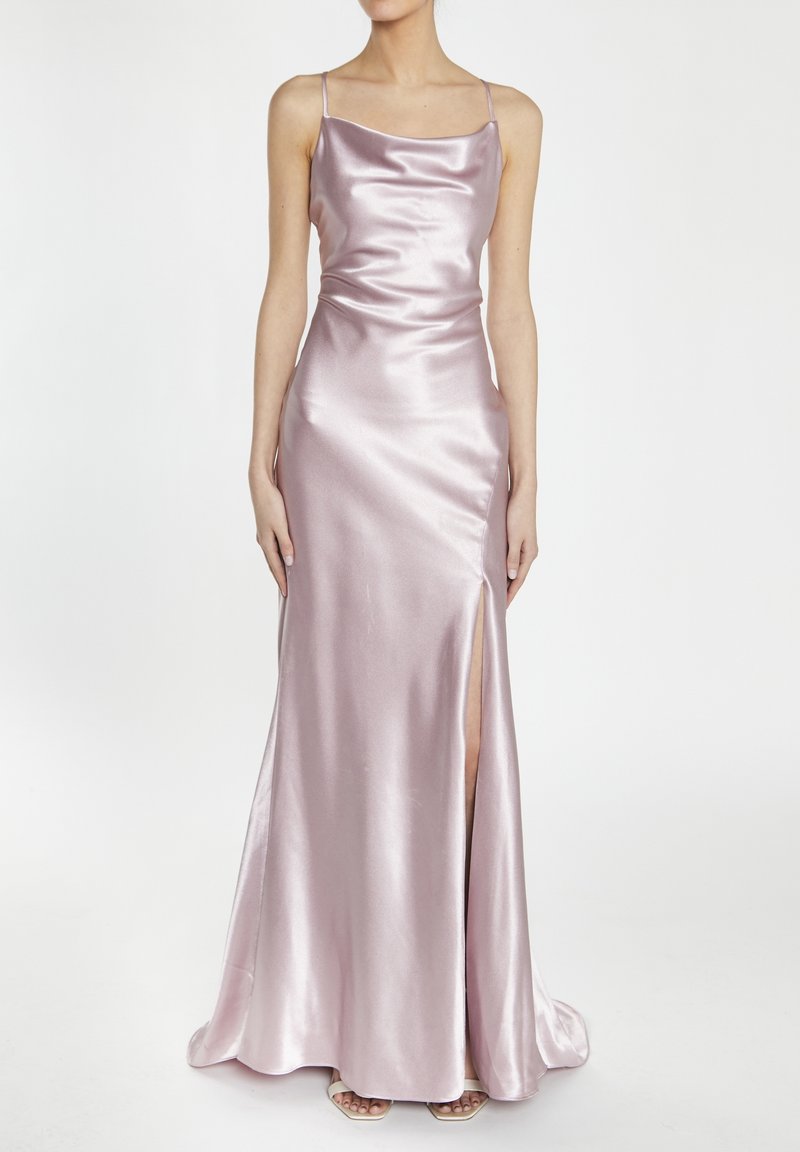 Femme portant une robe sans manches en satin rose clair avec de fines bretelles et une fente haute sur la cuisse, debout sur un fond blanc uni.