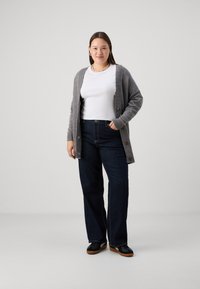 Cardigan gris con botones sobre una camisa blanca, combinado con pantalones vaqueros de pierna ancha azul oscuro. Zapatos negros con suelas marrones, con accesorios mínimos visibles.