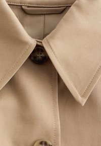 Close-up of beige collared shirt with visible stitched seams and dark round buttons on textured fabric.