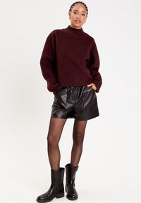 Pull en polaire bordeaux à col montant, associé à un short en cuir noir et des bottes noires hautes. Texture lisse et coupe décontractée.