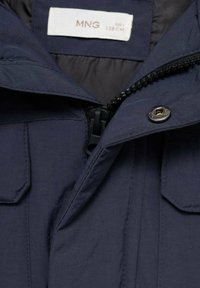 Detailný záber na golier námornícko-modrej bundy, ktorý zobrazuje zips, kovový cvok, vrecká s chlopňou a veľkostnú etiketu označenú ako MNG 128 CM.