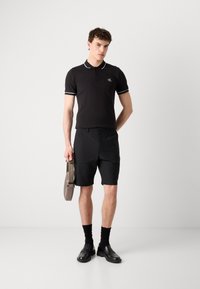 Svart polo-skjorte med hvit kant, kombinert med svarte shorts. Modellen holder en grå bag, iført svarte loafers og knelange sorte sokker.