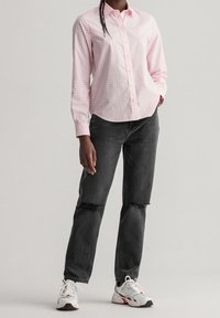Een persoon staat vol zelfvertrouwen in een roze geruit shirt, zwarte gescheurde jeans en witte sneakers, tegen een effen achtergrond.