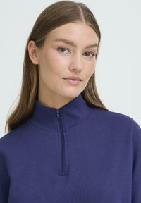 Mornarsko moder pulover z delno zadrgo in dolgimi rokavi. Mehka tekstura tkanine; brez vidnih vzorcev ali poudarkov.